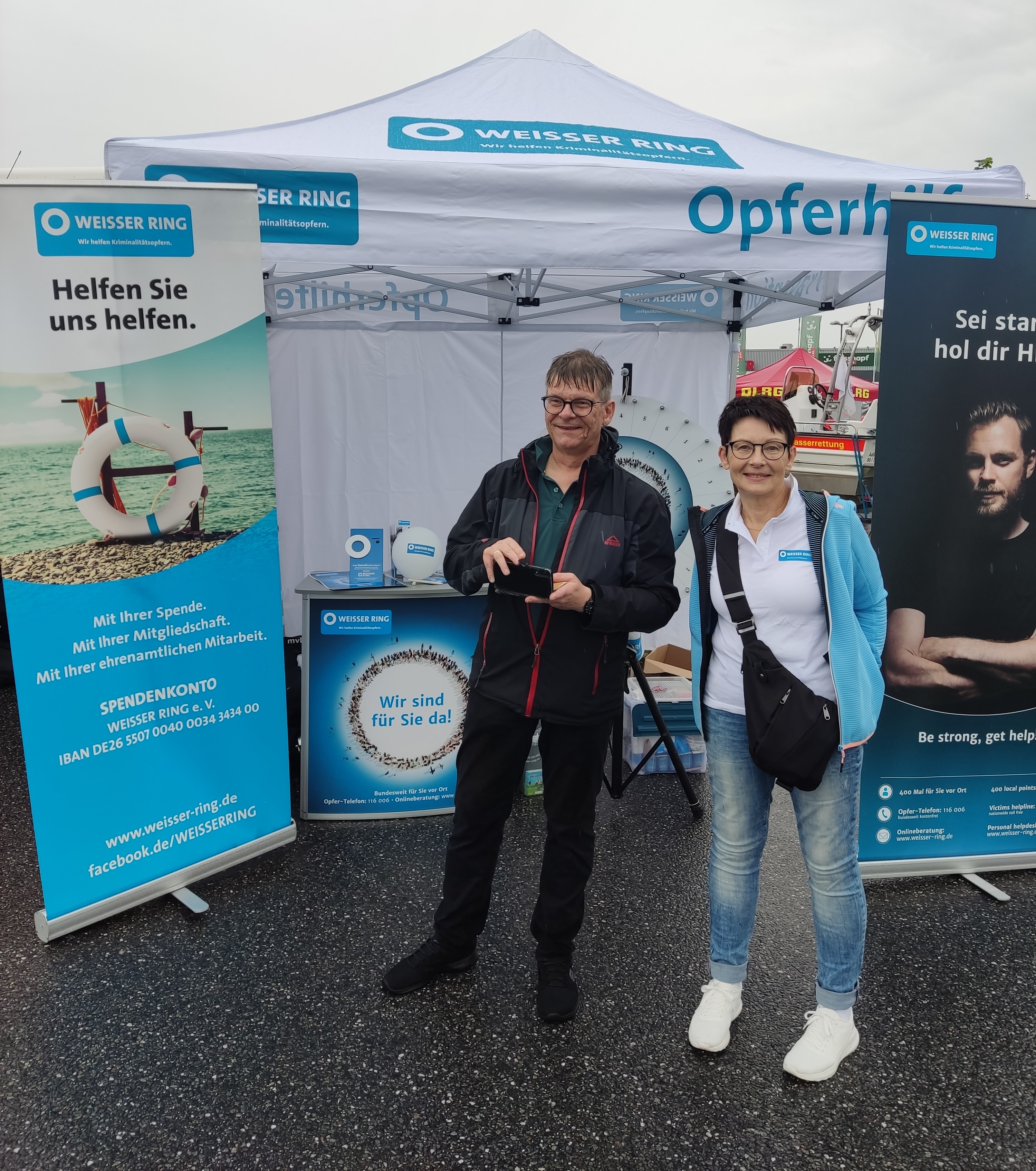2 Mitarbeiter vor dem Stand, eine Mitarbeiterin schaut lächelnd in die Kamera, der andere Mitarbeiter filmt mit seinem Handy etwas, was hinter der Kamera liegt und grinst