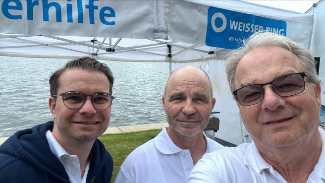 3 Mitarbeiter vor dem Stand, im Hintergrund ist das Logo vom Weissen Ring am Zelt zu sehen