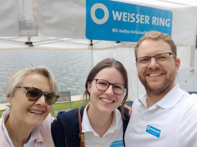3 Mitarbeiter vor dem Stand, im Hintergrund ist das Logo des Weissen Rings am Zelt zu sehen