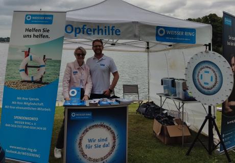 2 Mitarbeiter des Weissen Rings vor dem Stand, im Hintergrund der See und graue Wolken am Himmel